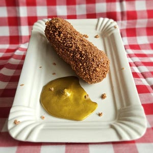 May include: A brown, breaded food item, possibly a croquette or similar, sits on a white, rectangular plate with a scalloped edge. A pool of golden sauce is next to it. The background is a red and white checkered tablecloth.