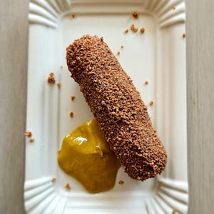 May include: A rectangular white paper plate holds a breaded, brown, cylindrical food item with a dollop of yellow mustard. The food item is covered in small, textured pieces. Scattered crumbs are on the plate.