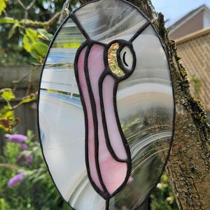 May include: Oval stained glass art piece with a pink and white design, outlined in black. The art piece is hanging from a silver chain and is set against a blurred background of greenery and a tree trunk. The design includes a gold accent.