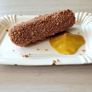 May include: A rectangular, white paper plate with a fluted edge holds a breaded, brown food item and a dollop of yellow sauce. The food item is covered in small, textured pieces. The plate sits on a light-colored wooden surface.