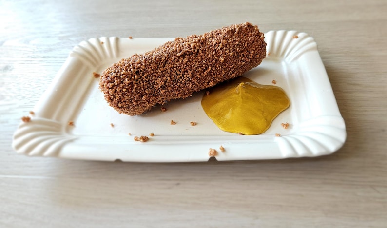 May include: A rectangular, white paper plate with a fluted edge holds a breaded, brown food item and a dollop of yellow sauce. The food item is covered in small, textured pieces. The plate sits on a light-colored wooden surface.