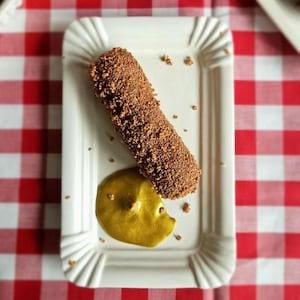 May include: A rectangular white plate with a fluted edge holds a chocolate-covered dessert and a dollop of yellow sauce. The dessert is a cylindrical shape, covered in a textured, dark brown coating. The plate sits on a red and white checkered tablecloth.