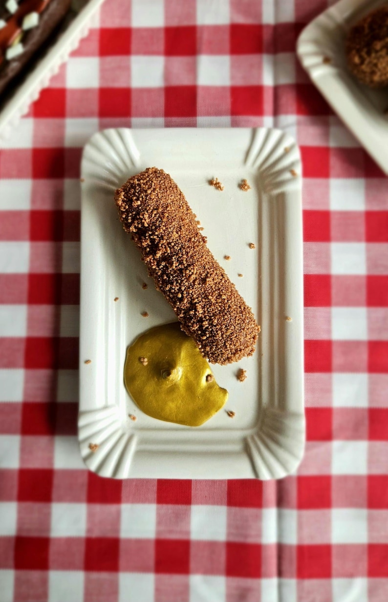 May include: A rectangular white plate with a fluted edge holds a chocolate-covered dessert and a dollop of yellow sauce. The dessert is a cylindrical shape, covered in a textured, dark brown coating. The plate sits on a red and white checkered tablecloth.