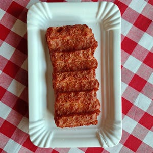 Puede incluir: Un plato rectangular de papel blanco contiene una pila de alimentos de color marrón rojizo. La comida parece estar frita o a la parrilla, con una superficie texturizada. El plato está sobre un mantel a cuadros rojos y blancos, evocando un picnic.