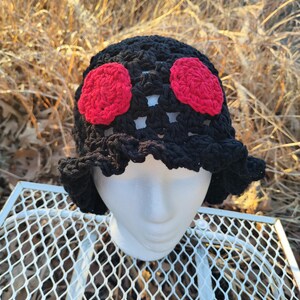 May include: A black crochet hat with two red flower accents. The hat has a ruffled brim and is displayed on a white mannequin head. The hat is sitting on a white metal table with a diamond pattern.