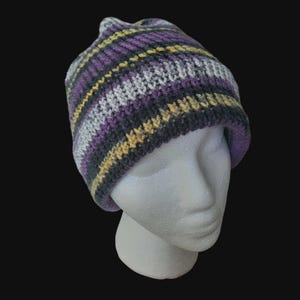 May include: A knitted beanie hat with horizontal stripes of purple, gray, yellow, and black. The hat is displayed on a white mannequin head against a black background. The hat is a winter accessory.