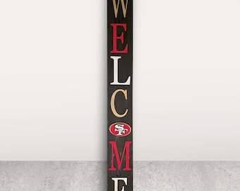 Puede incluir: Un letrero de bienvenida de madera negra con la palabra "WELCOME" en colores alternos. Las letras son blancas, rojas y doradas, con el logotipo de los 49ers de San Francisco. El letrero está diseñado para uso en exteriores.