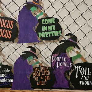 May include: Halloween-themed wooden decoration featuring witch cutouts with phrases like "Hocus Pocus" and "Toil and Trouble." The witches are dressed in purple robes and black hats, holding green-handled brooms. The decoration is set against a black net background.