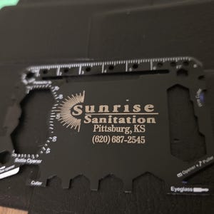 Può includere: Una scheda multi-strumento nera con vari ritagli per diverse funzioni, tra cui un apribottiglie, un taglierino e un tronchese. La scheda presenta il testo "Sunrise Sanitation Pittsburg, KS (620) 687-2545" in lettere argentate.