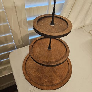 May include: A three-tiered wooden serving stand with a dark brown finish. The stand has three circular trays of varying sizes, connected by a central black metal rod. The top handle is a decorative, twisted design.