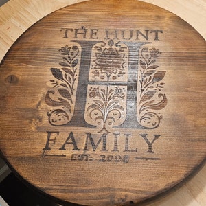Puede incluir: Un letrero redondo de madera con el texto "The Hunt Family Est. 2008" en una fuente marrón oscura. La letra "H" es una inicial grande con un diseño floral.