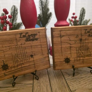 May include: Two wooden plaques with the text "LaCynge Kansas" and a map design are displayed on decorative stands. The plaques are made of light brown wood and feature a compass rose design. The background includes red vases and greenery.