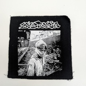 May include: Black square patch with a black and white image of a person wearing goggles and a head covering. The patch has the word "Astoria" in white, stylized lettering at the top.