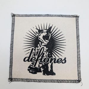May include: Square, off-white patch with a black illustration of a roaring cat with sunburst rays. The word "deftones" is written in a stylized font below the cat. The patch has a black stitched border.