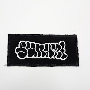 May include: A black rectangular patch with white puffy lettering that reads "SURFACE". The patch has a black border with visible stitching. The background is plain white.