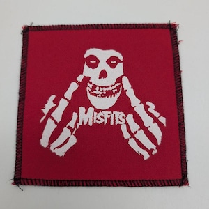 May include: Square red patch featuring a white skull with skeletal hands and the word "MISFITS" in white. The patch has a black stitched border.