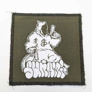 May include: An olive green square patch with a black stitched border. The patch features a white graphic of a hooded figure holding a gun, with a dollar sign on the chest. The word "SHRASS" is written in white graffiti-style lettering.