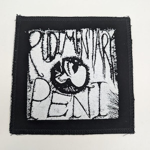 May include: Black square patch with white text that reads "RUDIMENTARY PENI" with a graphic in the center. The patch has a black border with a stitched edge. The text and graphic are in a distressed style.