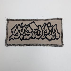 May include: A rectangular patch with a tan background and black embroidered text. The text is stylized in a graffiti art style. The patch has a black stitched border.