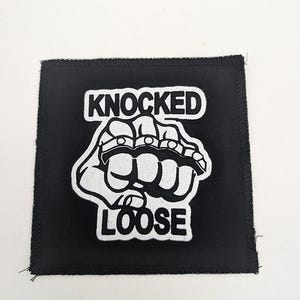 May include: Black square patch with white embroidered design. The design features a fist wearing brass knuckles, with the words "KNOCKED LOOSE" above and below the fist in a bold, sans-serif font. The patch has a stitched border.