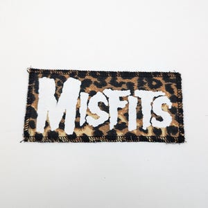 May include: A rectangular patch with the word "MISFITS" in white, distressed lettering. The patch has a leopard print background and a black stitched border. The patch is likely for clothing or accessories.