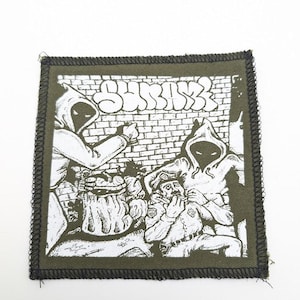 May include: A square patch with a black border and a graphic illustration in white. The illustration depicts two figures in hooded cloaks, a brick wall, and a person in a police uniform. The word "SHRINK" is written in graffiti style.
