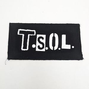 May include: Black rectangular patch with white stencil-style lettering spelling out "T.S.O.L.". The patch has a raw edge and is likely made of fabric. The letters are bold and stand out against the black background.