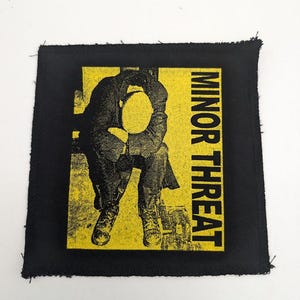 May include: Black square patch with a yellow rectangle featuring a black silhouette of a person sitting with their head in their hands. The words "MINOR THREAT" are printed vertically in black on the right side of the yellow rectangle.