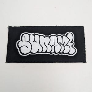 May include: Black rectangular patch with white embroidered text that reads "SURFSS" in a bubble-style font. The patch has a black border with visible stitching around the edges. The background is plain white.