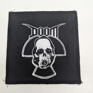 May include: Square black patch with a white graphic of a skull wearing a gas mask, with the word "DOOM" above it. The patch has a stitched border.