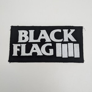 May include: Black rectangular patch with the white text "BLACK FLAG" and three vertical white lines. The patch is made of a textured fabric and is likely an iron-on or sew-on patch.