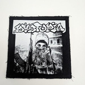 May include: Black and white square patch featuring the word "Dystopia" in stylized lettering above a grayscale image of a person holding a rifle. The patch has a black border.