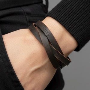 May include: A dark brown leather wrap bracelet featuring a smooth finish and a layered design. The leather band is embossed with the name Riley in a simple, clean font. The bracelet is secured around the wrist, showing a flexible construction.