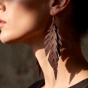 Op de afbeelding: Close-up van een paar bruinleren oorhangers in de vorm van een veer. De oorbellen zijn lang en bungelen, met ingewikkelde uitgesneden details. Ze zijn opgehangen aan goudkleurige haken. De achtergrond is neutraal en wazig.