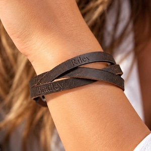 May include: A dark brown leather wrap bracelet featuring two distinct leather strands that cross over each other. The leather is engraved with the names Riley and Bradley in a simple, clear font, creating a personalized accessory.
