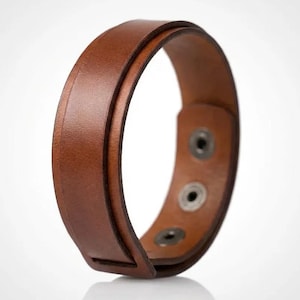 May include: A brown leather bracelet featuring a smooth, wide band design with a layered appearance. The bracelet includes three visible metal snap fasteners on the inner side, allowing for adjustable closure and a secure fit around the wrist.