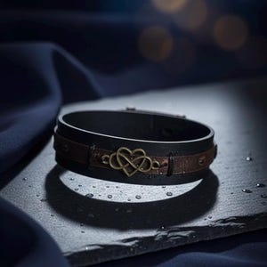 Puede incluir: Una pulsera de cuero negro con una banda de cuero marr&oacute;n y un dije de coraz&oacute;n infinito de color dorado. La pulsera se exhibe sobre una superficie oscura con gotas de agua.