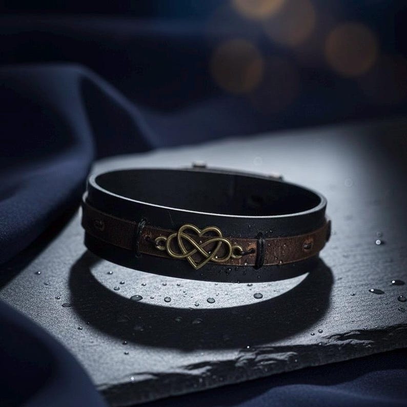 Puede incluir: Una pulsera de cuero negro con una banda de cuero marr&oacute;n y un dije de coraz&oacute;n infinito de color dorado. La pulsera se exhibe sobre una superficie oscura con gotas de agua.