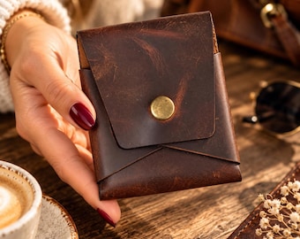 May include: A dark brown leather card holder with a folded envelope-style flap and a single circular brass-colored metal snap button closure. The leather features a distressed, textured surface with visible grain and natural markings.