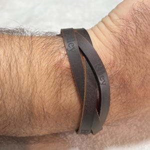 May include: A dark brown leather wrap bracelet featuring multiple strands that overlap. The leather surface is embossed with the names Bradley and Riley in a clear, sans-serif font. The bracelet is designed to wrap around the wrist multiple times.