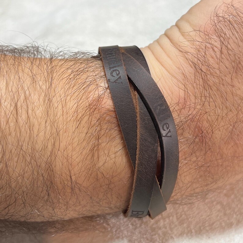 May include: A dark brown leather wrap bracelet featuring multiple strands that overlap. The leather surface is embossed with the names Bradley and Riley in a clear, sans-serif font. The bracelet is designed to wrap around the wrist multiple times.