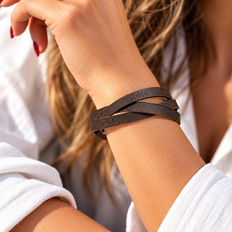 May include: A dark brown leather wrap bracelet featuring two distinct leather strands that cross over each other. The leather is engraved with the names Riley and Bradley in a simple, clear font, creating a personalized accessory.