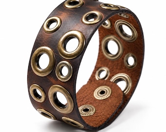 May include: A wide dark brown leather cuff bracelet featuring multiple brass-toned metal grommets of varying sizes scattered across the surface. The bracelet is secured with two brass-toned metal snap fasteners on the inner leather strap.