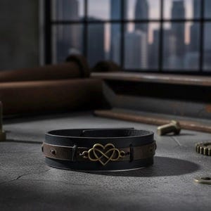 Puede incluir: Una pulsera de cuero negro con una banda de cuero marr&oacute;n y un dije de coraz&oacute;n infinito de color dorado. La pulsera est&aacute; sobre una superficie gris con un fondo borroso de un paisaje urbano y herramientas industriales.