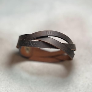 May include: A dark brown leather bracelet featuring multiple intertwined straps. The leather surface is engraved with the names Riley and Bradley in a clear, dark font. The bracelet is secured with a metallic snap button fastener.