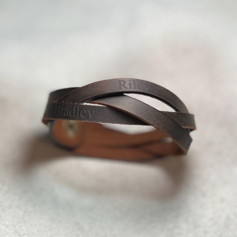 May include: A dark brown leather bracelet featuring multiple intertwined straps. The leather surface is engraved with the names Riley and Bradley in a clear, dark font. The bracelet is secured with a metallic snap button fastener.