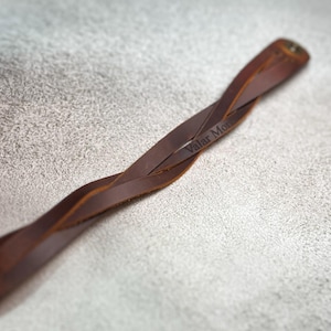 May include: A brown leather bracelet featuring a twisted design and a metal snap closure. The leather strap is engraved with the text Valar Morghulis, visible along the surface of the twisted band, showcasing a smooth finish and natural texture.