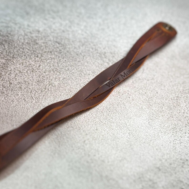 May include: A brown leather bracelet featuring a twisted design and a metal snap closure. The leather strap is engraved with the text Valar Morghulis, visible along the surface of the twisted band, showcasing a smooth finish and natural texture.