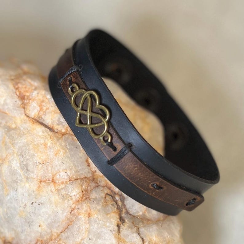 Puede incluir: Pulsera de cuero negro con un acento de cuero marr&oacute;n y un dise&ntilde;o en forma de coraz&oacute;n de color dorado. La pulsera tiene un cierre de hebilla y mide aproximadamente 2,5 cm de ancho.