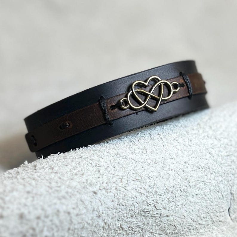 Puede incluir: Una pulsera de cuero negra y marr&oacute;n con un s&iacute;mbolo de infinito en forma de coraz&oacute;n de color dorado. La pulsera est&aacute; hecha de dos capas de cuero, con la capa superior en negro y la capa inferior en marr&oacute;n. La pulsera mide aproximadamente 3,8 cm de ancho.